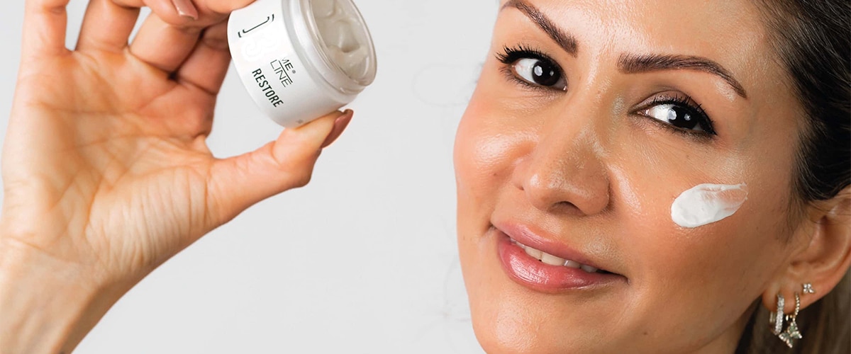 Vrouw glimlacht terwijl ze een potje huidcrème omhooghoudt en een witte streep crème op haar wang dept, illustrerend hoe je melasma in de zomer kunt verzorgen.